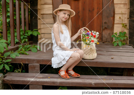 Playful tween girl sitting on doorstep of country house with basket of wildflowers 87518036