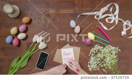 Top view of a table with items to create a composition for Easter. A woman's hand writes with a red marker: Easter menu. Christ is risen. Church holiday-Easter Top view of a table with items to create a composition for Easter. A woman's hand writes with a red marker: Easter menu. Christ is risen. Church holiday-Easter 87520729