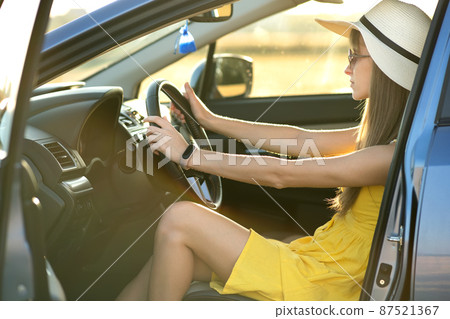 Young driver woman in yellow dress and straw hat sitting behind steering wheel driving a car. Summer vacation and travel concept. 87521367