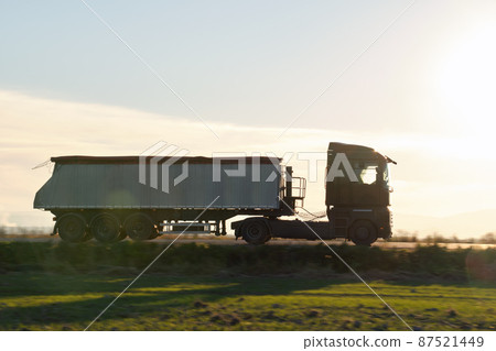 Semi-truck with tipping cargo trailer transporting sand from quarry driving on highway hauling goods in evening. Delivery transportation and logistics concept Semi-truck with tipping cargo trailer transporting sand from quarry driving on highway hauling goods in evening. Delivery transportation and logistics concept 87521449