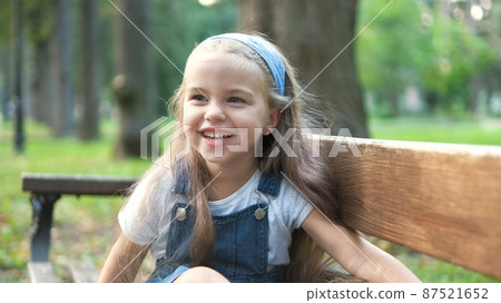 Little happy child girl sitting on a bench smiling happily in summer park. Little happy child girl sitting on a bench smiling happily in summer park. 87521652