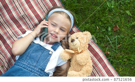 Little cute child girl laying on a blanket on green lawn in summer outdoors with her teddy bear toy talking on mobile phone. Little cute child girl laying on a blanket on green lawn in summer outdoors with her teddy bear toy talking on mobile phone. 87521656