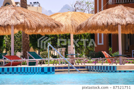 Empty deck chairs under straw shade umbrellas on swimming pool side in tropical resort. Summer vacations and getaway concept Empty deck chairs under straw shade umbrellas on swimming pool side in tropical resort. Summer vacations and getaway concept 87521751