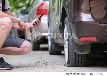 Driver calling road service for assistance having vehicle trouble with punctured flat tire on car parked on roadside Driver calling road service for assistance having vehicle trouble with punctured flat tire on car parked on roadside 87521760