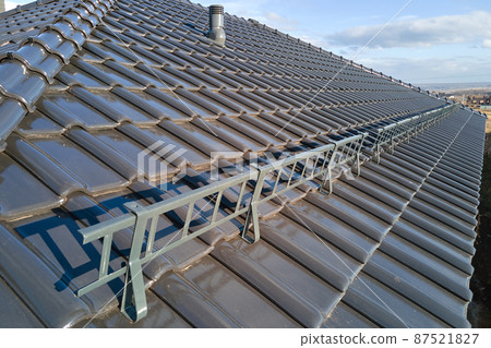 Closeup snow guard for safety in winter on house roof top covered with ceramic shingles. Tiled covering of building 87521827