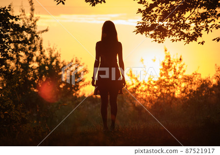Back view of young woman in summer dress walking alone through evening dark forest 87521917