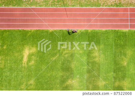 Aerial view of small figure of worker cutting green grass with mowing mashine on football stadium field with red running tracks in summer. Aerial view of small figure of worker cutting green grass with mowing mashine on football stadium field with red running tracks in summer. 87522008