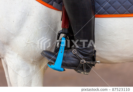 The foot of the rider, sitting on a white horse, in a black boot with a spur, rests on a safe plastic stirrup. Close-up. Equestrian competition show. Thoroughbred beautiful stallion 87523110
