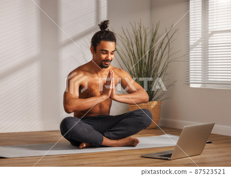 Young friendly ethnic man in casual wear sitting in lotus pose in front of laptop at home Young friendly ethnic man in casual wear sitting in lotus pose in front of laptop at home 87523521