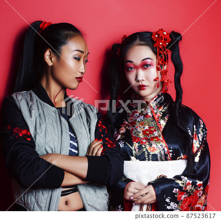 two pretty geisha friends: modern asian woman and traditional wearing kimono posing cheerful on red background 87523617