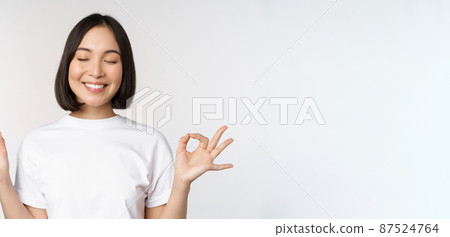 Portrait of young asian woman meditating, smiling pleased and practice yoga, close eyes and meditate, breathing calm, standing over white background 87524764