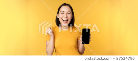 Excited asian woman, showing smartphone app and triumphing, celebrating on mobile phone, standing over yellow background 87524796