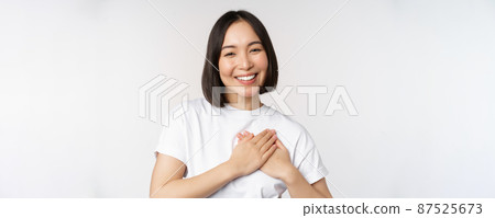 Close up of smiling korean woman holding hands on heart, care and love concept, feel affection, tenderness or heartwarming feeling, standing over white background Close up of smiling korean woman holding hands on heart, care and love concept, feel affection, tenderness or heartwarming feeling, standing over white background 87525673