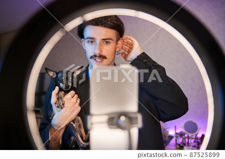 Man standing in front of the smartphone and posing to the camera with his lovely little dog Man standing in front of the smartphone and posing to the camera with his lovely little dog 87525899