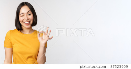 Beautiful young woman showing okay sign, smiling pleased, recommending smth, approve, like product, standing in yellow tshirt over white background 87525909