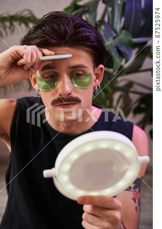 Serious man using facial gua sha jade board while looking at the mirror at his reflection 87525974