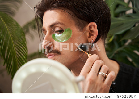 Serious young guy is having anti-wrinkle treatment while looking at the mirror and massaging Serious young guy is having anti-wrinkle treatment while looking at the mirror and massaging 87525997