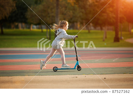 Portrait of active little toddler girl riding scooter on road in park outdoors on summer day. Seasonal child activity sport. Healthy childhood lifestyle Portrait of active little toddler girl riding scooter on road in park outdoors on summer day. Seasonal child activity sport. Healthy childhood lifestyle 87527162