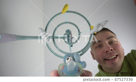 A young father gives a pacifier to a newborn baby lying in a cradle with a baby mobile with toys. First-person view from the bottom up. 87527330