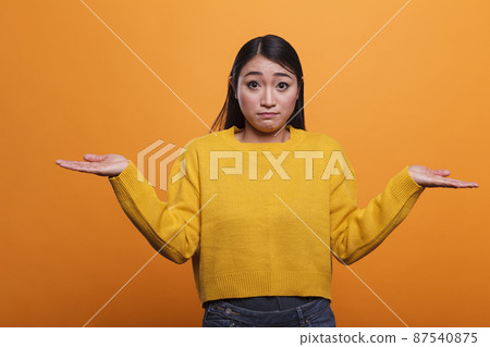 Confused uncertain asian woman shruging unsurely while wearing yellow sweater on orange background. Confused uncertain asian woman shruging unsurely while wearing yellow sweater on orange background. 87540875