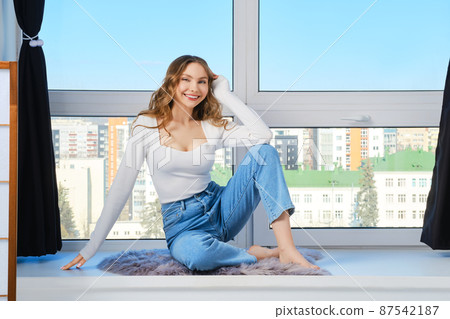 Barefoot young woman sits on windowsill leaning on her knee Barefoot young woman sits on windowsill leaning on her knee 87542187