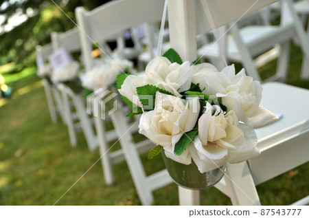 White Chairs Adorned with White Fabric Rose Bouquets Await Guests at a Garden Wedding 87543777