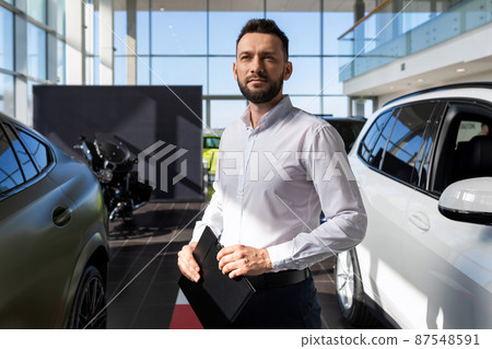 automobile center specialist against the backdrop of a car dealership with a folder in his hands automobile center specialist against the backdrop of a car dealership with a folder in his hands 87548591