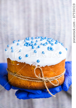 Hands in blue gloves hold Easter cake with white topping and blue sprinkles. Woman holding traditional Russian Easter cake. Homemade pie for spring holiday celebration Hands in blue gloves hold Easter cake with white topping and blue sprinkles. Woman holding traditional Russian Easter cake. Homemade pie for spring holiday celebration 87550319