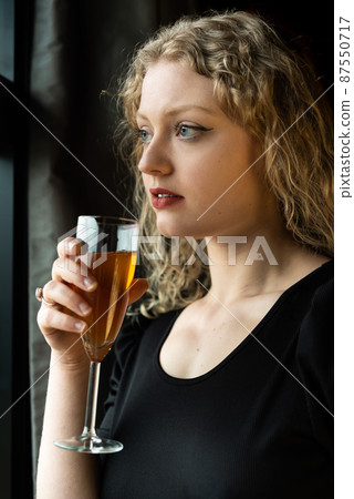 Window portrait of a 21 year old white blonde woman with curling hair holding a glass of champaign Window portrait of a 21 year old white blonde woman with curling hair holding a glass of champaign 87550717