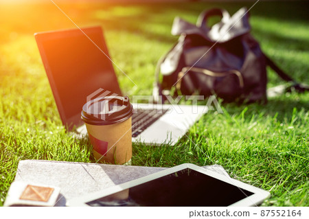 Laptop computer on green grass with coffee cup, bag and tablet in outdoor park Laptop computer on green grass with coffee cup, bag and tablet in outdoor park 87552164