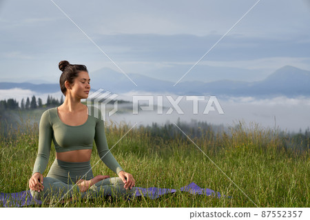 Young woman with dark hair practising meditation in lotus position with beautiful mountains on background. Concentration and relaxation concept. Young woman with dark hair practising meditation in lotus position with beautiful mountains on background. Concentration and relaxation concept. 87552357