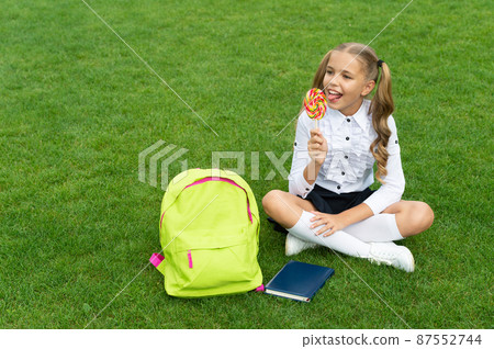 Happy girl child in uniform with school bag and book enjoy eating tasty lollipop sitting on grass Happy girl child in uniform with school bag and book enjoy eating tasty lollipop sitting on grass 87552744