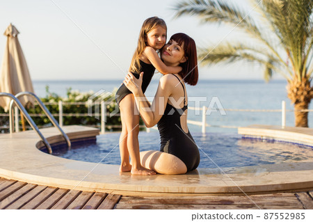 Mother and daughter in black bikini are sitting at the pool edge and enjoying summer vacation time 87552985
