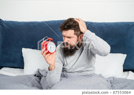 Perplexed guy scratching head holding alarm clock being in bed in morning, oversleep 87553064