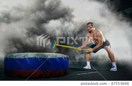 Strong man with Ukrainian flag on cheeck hitting with hammer giant wheel with Russian flag on it isolated on fog, smoke 87553698