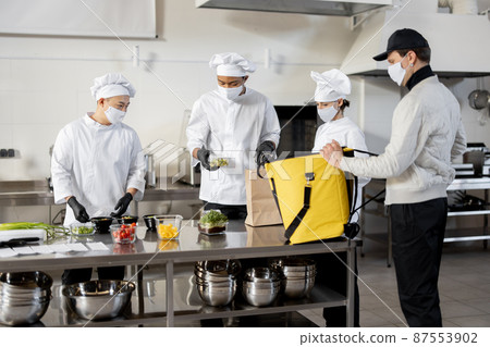 Courier waiting for an order for delivery in the kitchen with chefs preparing takeaway food 87553902