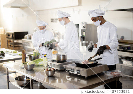 Multiracial team of cooks in uniform and face masks cooking meals for a restaurant in the kitchen Multiracial team of cooks in uniform and face masks cooking meals for a restaurant in the kitchen 87553921