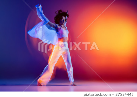 Street dancing. Image of flexible young girl, hip-hop dancer in white outfit dancing hip hop isolated on blue background in yellow neon light. Youth culture, style and fashion, action. 87554445