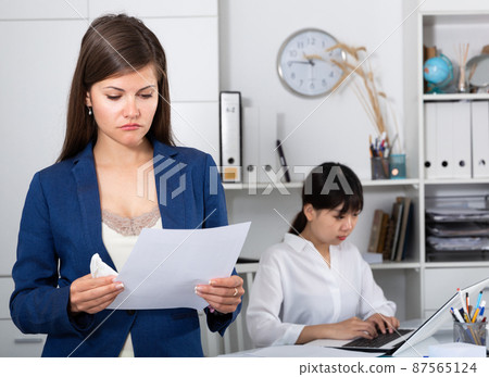 Business woman crying standing in office with working colleague behind Business woman crying standing in office with working colleague behind 87565124