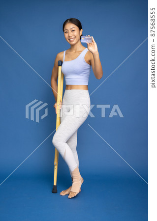 Portrait of a Asian woman wearing yoga or exercise clothes using a crutch and showing credit card on hand isolated on blue background, Foot injury and Exercise accident concept 87568555