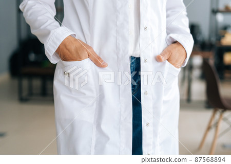 Close-up cropped shot of unrecognizable female doctor in white uniform holding hands in pockets standing in hospital office. Practitioner woman posing at workplace. Concept of medical work. 87568894