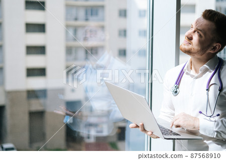 Blinking from sun male doctor in white uniform with stethoscope using laptop holding with one hand, standing near window in sunny day. Concept of modern medicine and healthcare. 87568910