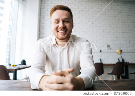 Close-up portrait of handsome happy young business man sitting at desk in modern office room and smiling looking at camera. Concept of successful worker, career advance and opportunity. Close-up portrait of handsome happy young business man sitting at desk in modern office room and smiling looking at camera. Concept of successful worker, career advance and opportunity. 87569003