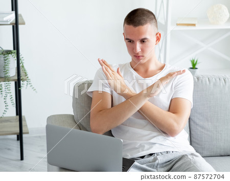 Stop gesture. Protesting man. Disagreed expression. Handsome strict casual guy crossing hands showing on laptop sitting sofa in light room interior. 87572704