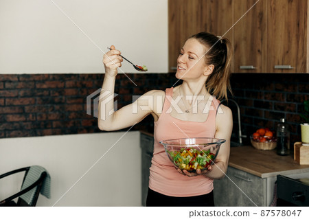 Portrait of an attractive woman holding a salad bowl and looking at the camera. Beautiful athletic girl in sportswear eating healthy salad after workout at home Portrait of an attractive woman holding a salad bowl and looking at the camera. Beautiful athletic girl in sportswear eating healthy salad after workout at home 87578047
