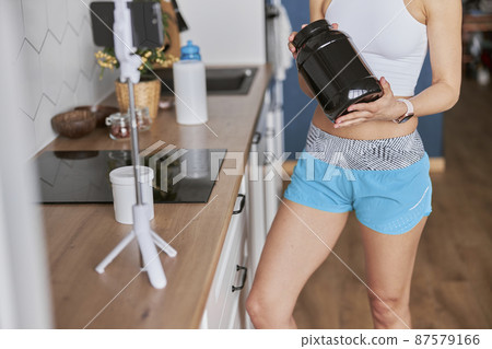 Lady trainer shows black jar of protein supplement to followers in light kitchen closeup Lady trainer shows black jar of protein supplement to followers in light kitchen closeup 87579166