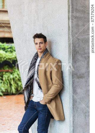 Portrait of stylish handsome young man standing outdoors and leaning on wall. Man wearing jacket and scarf Portrait of stylish handsome young man standing outdoors and leaning on wall. Man wearing jacket and scarf 87579204