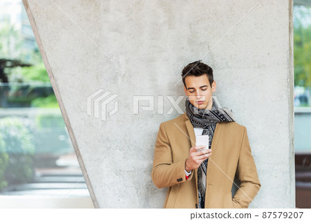 Businessman texting on mobile phone against wall Businessman texting on mobile phone against wall 87579207