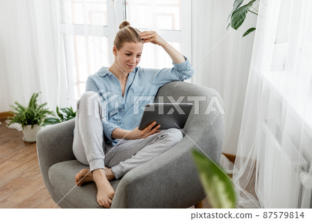 Beautiful young woman in cozy home clothes communicates with work colleagues on a tablet at home in the living room 87579814