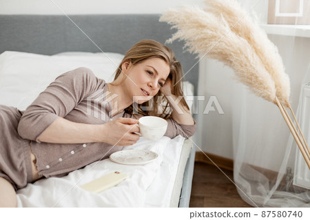 Young beautiful woman in light pajamas enjoys morning in bed with coffee in cozy bedroom. Morning rituals Young beautiful woman in light pajamas enjoys morning in bed with coffee in cozy bedroom. Morning rituals 87580740